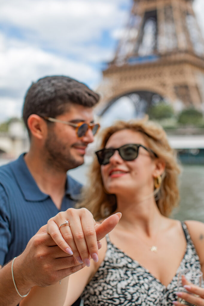 Close-up photo of a couple holding hands with a focus on the engagement ring on the lady's hand. The Eiffel tower is visible in the blurry background