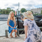 A mother takes a smartphone photo of her young blond daughter, who is sitting on the river bank in front of a boat