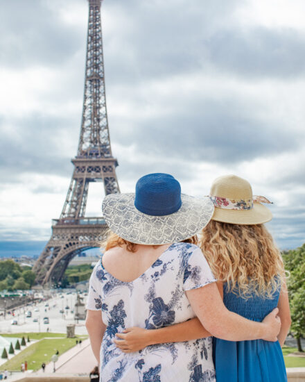 A mother and daughter in wide-bream hats with their backs towards the camera are embracing each other as they admire the Eiffel Tower