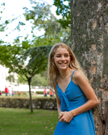 A blond girl in a blue dress is smiling as she poses next to a tree in Trocadero gardens in Paris