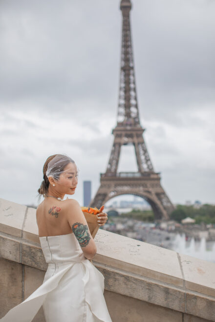 An Asian bride with tattoes and a bouquet of orange roses is posing in front of the Eiffel Tower. Radina Dianova - Paris based photographer