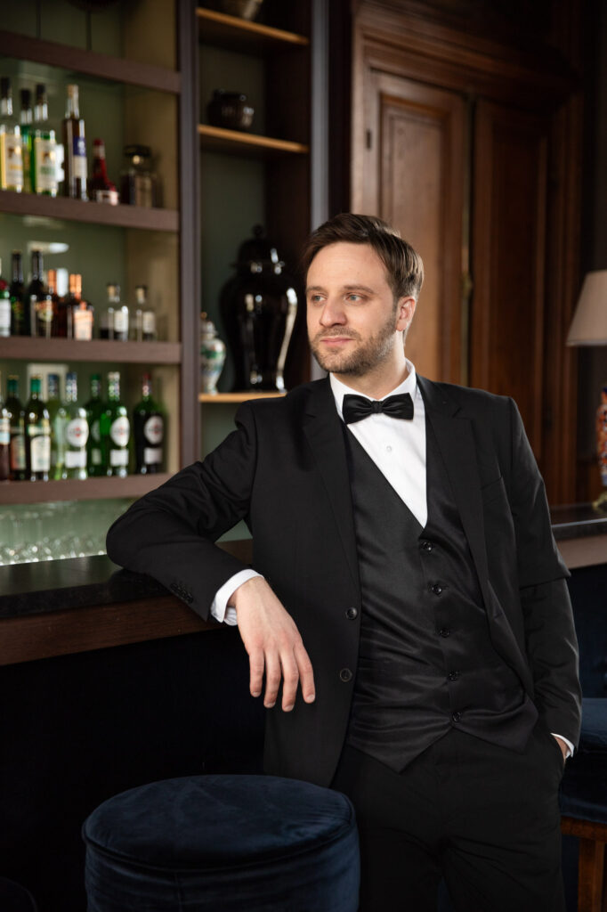 Groom portrait in black tuxedo leaning against bar inside French château wedding venue